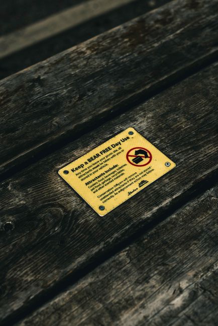A close-up image of a rectangular yellow metal sign affixed with four screws to a weathered, dark brown wooden surface, likely part of outdoor furniture or a structural element. The sign displays black text and a red and black pictogram. The text advises to keep the area bear free and to use the designated bin for disposal, instructing not to feed the bears and indicating attraction issues. The background appears blurred, suggesting an outdoor environment, with natural lighting highlighting the textured, aged wood grain and the contrast of the bright sign. The scene relates to outdoor waste management or local guidelines for waste disposal and animal safety, reflecting the importance of appropriate rubbish handling in communal or natural settings, supporting alternative disposal practices and waste awareness.