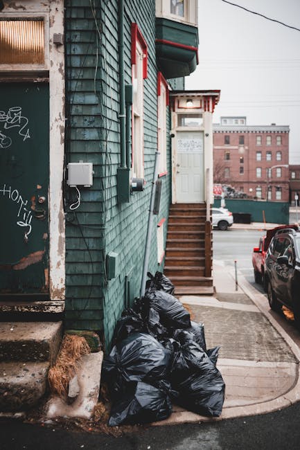 A cluster of black trash bags filled with rubbish is piled against the green exterior wall of a building on a city street. The building features weathered, dark green wooden siding with visible signs of wear and graffiti on a nearby door frame. A small window with a broken or smeared glass panel is situated above the bags, and several utility boxes and cables are mounted on the wall adjacent to the trash. To the right, a set of brown wooden stairs with a white door at the top leads to an entrance, and a small canopy extends above the door. The sidewalk is concrete, and the area appears to be in a busy urban environment with parked cars, including a black vehicle, visible along the street in the background. The scene suggests a typical independent rubbish disposal method, with rubbish bags positioned outside a residential or commercial building, reflecting an aspect of private waste handling often seen in urban settings. The lighting indicates an overcast day, with diffused light softly illuminating the scene.