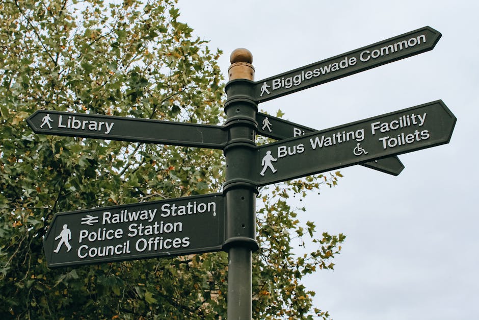 A black multi-directional street signpost features seven rectangular signs pointing in various directions against a background of green foliage and overcast sky. The signs are labeled with white text indicating locations such as 'Biggleswade Common', 'Library', 'Railway Station', 'Police Station', 'Council Offices', 'Bus Waiting Facility', and 'Toilets'. The 'Railway Station' sign includes an icon of a train, while the 'Toilets' sign has a wheelchair accessibility symbol. The signs have a matte finish, with some showing slight weathering, and are mounted on a central dark green pole with a rounded top. The scene suggests an urban or suburban environment near public amenities, with the signpost serving pedestrians navigating around community facilities. This image aligns with the context of rubbish collection and alternative waste handling by highlighting the importance of local infrastructure and directional signage within residential or town areas, referencing community services and public spaces that may be serviced by local rubbish removal companies like our firm, ensuring efficient waste collection from various public facilities.