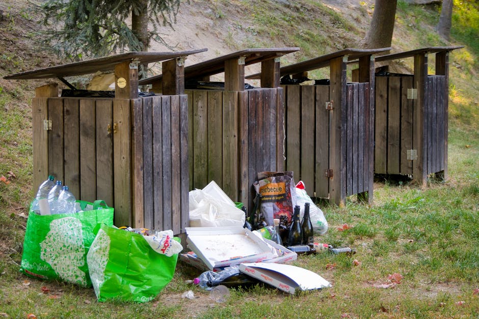 A series of four wooden outdoor rubbish bins with slanted, overhanging roofs are positioned on a grassy area near a slope with trees in the background. The bins are constructed from vertical wooden planks with visible hinges and padlocks on some. In front of the bins, there is an accumulation of waste, including a large green plastic bag filled with bottles and other refuse, a paper pizza box, and several clear plastic bottles, two dark glass bottles, and a few smaller plastic containers. The waste appears to be loosely scattered on the grass, indicating a collection or disposal activity. The scene is lit by natural daylight, with shadows cast by nearby trees, creating a calm outdoor setting. This image illustrates a typical outdoor rubbish disposal area, which could be indicative of private waste handling or alternative rubbish collection practices often seen in non-urban environments as part of a waste management service such as those offered by rubbish collection providers like rubbishcollectionbelsizepark.co.uk.