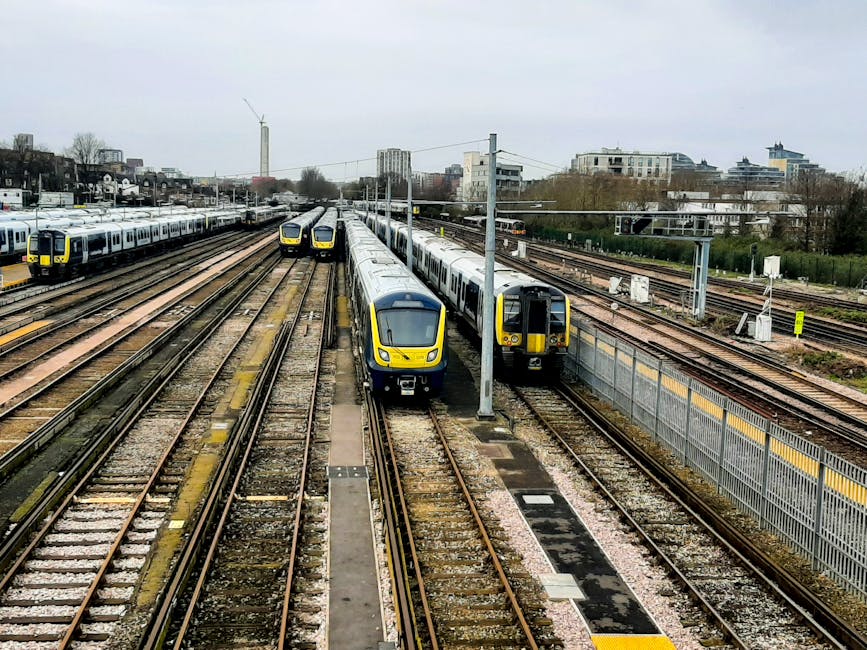 A view of a railway yard with multiple train tracks running parallel across the scene. Two modern passenger trains, predominantly silver with yellow front panels, are positioned on adjacent tracks towards the foreground. The train on the left has a sleek, curved front window, while the one on the right appears slightly older with a darker front design. To the left, several stationary trains are visible, mostly with white and blue exterior coloring, aligned along additional tracks. The tracks are made of metal rails mounted on wooden sleepers, with gravel ballast in between, showing signs of weathering and moss growth. Overhead electrical lines and poles stretch across the scene, supported by metal masts that run parallel to the tracks. In the background, a mixture of mid-rise residential and commercial buildings can be seen, some with balconies or rooftop structures. The sky is overcast with grey clouds, giving the scene a muted, industrial atmosphere. The entire scene appears to be part of a rail depot or maintenance yard, with the context related to private or alternative railway operations, similar in function to independent rubbish collection but in the transport sector, as observed in the rail infrastructure layout.